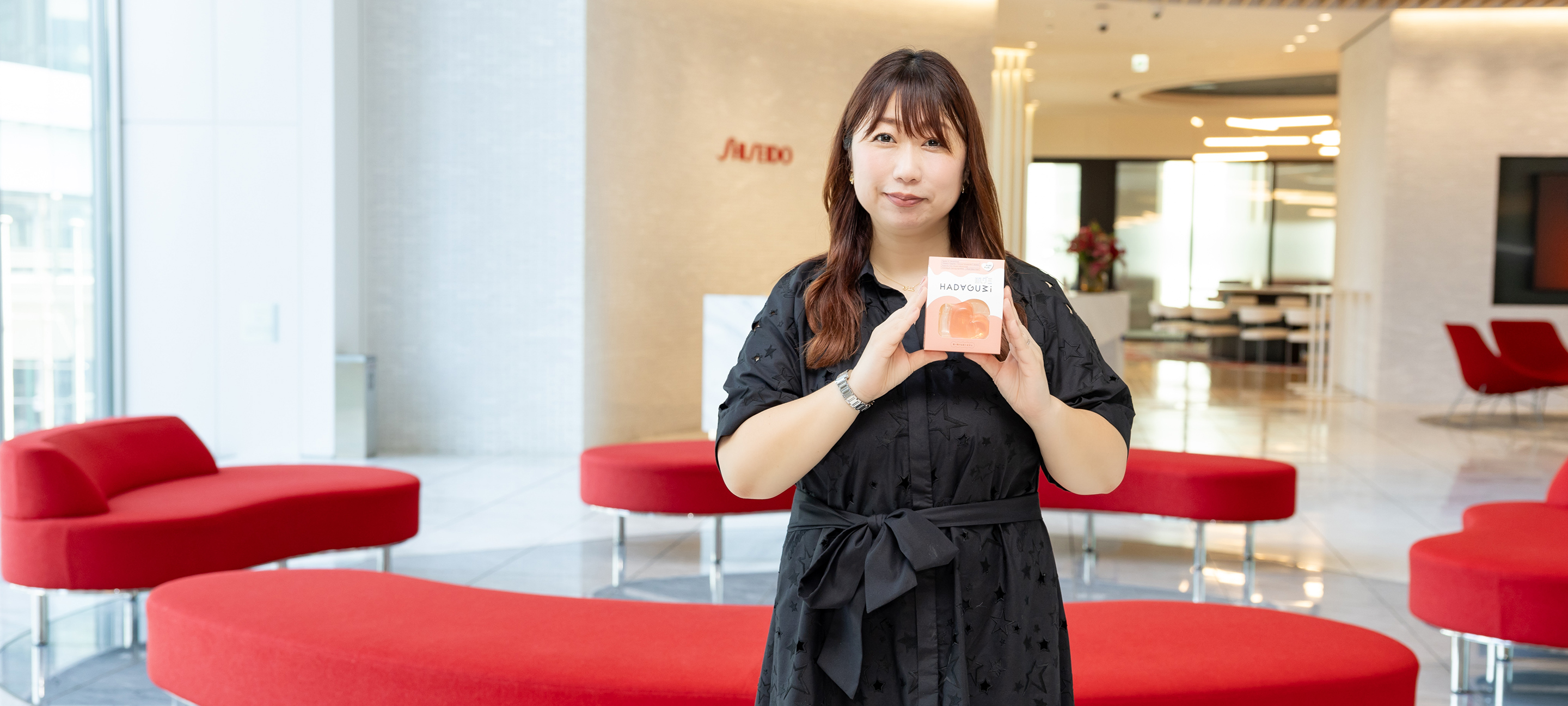 資生堂社員の釣谷が、資生堂汐留オフィスのロビーを背景に、洗顔料である肌グミパッケージを手にして笑顔で立っている