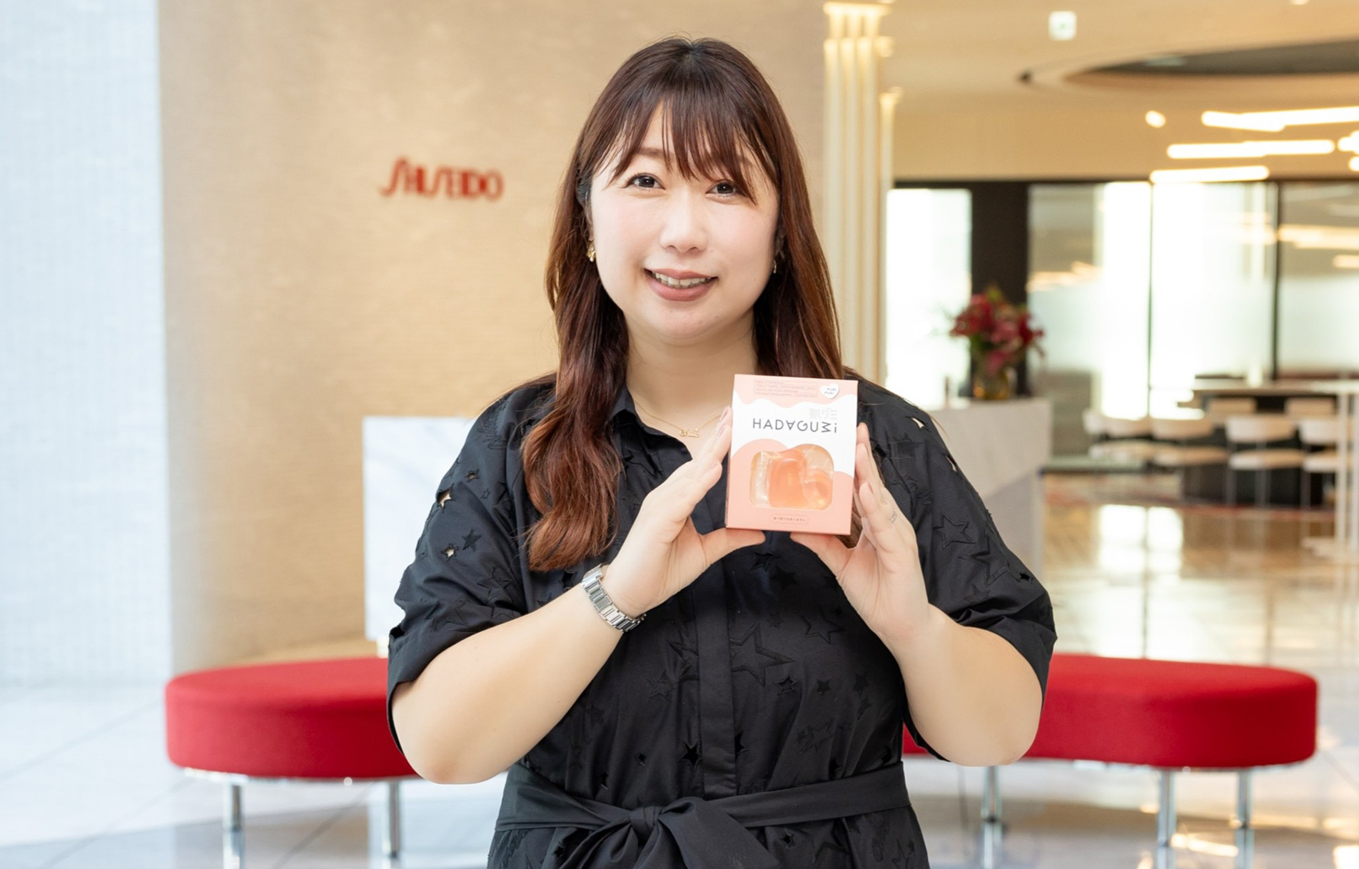 資生堂社員の釣谷が、資生堂汐留オフィスのロビーを背景に、洗顔料である肌グミパッケージを手にして笑顔で立っている